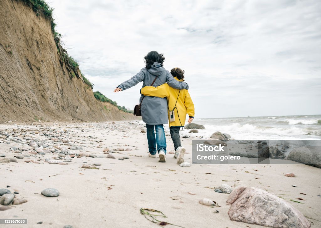 little girl in yellow raincoat walking embraced with mother on stone beach rear view on little girl in yellow raincoat walking embraced with mother at beach of the baltic sea Vacations Stock Photo