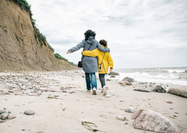 little girl in yellow raincoat walking embraced with mother on stone beach stock photo