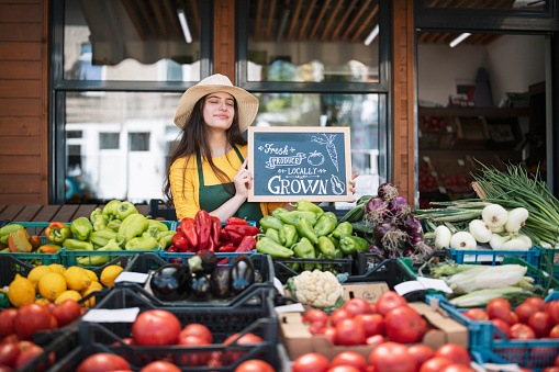 Portrait of a young female greengrocer in front of her store holding a sign and smiling
