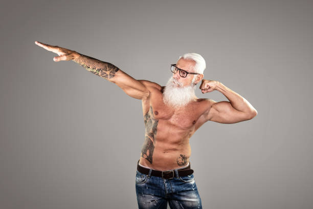 Happy muscular senior man showing his muscular body and tattoos Happy muscular senior man showing his muscular body and tattoos while posing in studio over grey background. Age is just a number. A lot of copy space. senior bodybuilders stock pictures, royalty-free photos & images