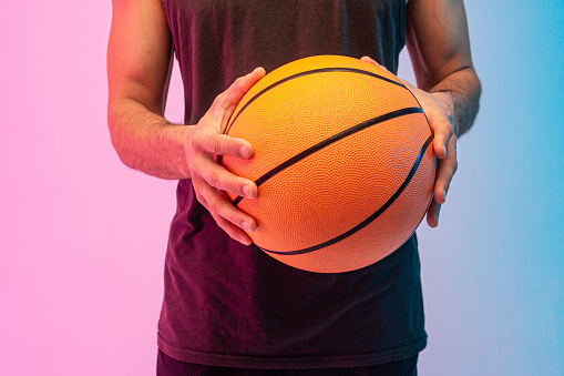 Close up image of man hold basketball ball in hands. Guy wear tank top. Isolated on blue and pink background. Studio shoot