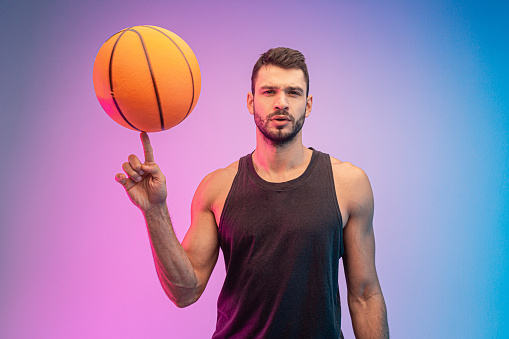 Confident sportsman spinning basketball ball on finger. Young bearded european basketball player looking at camera. Isolated on blue and pink background. Studio shoot. Copy space