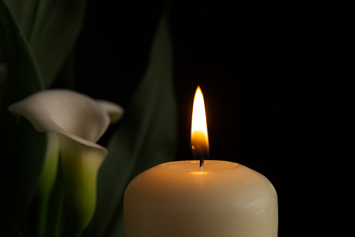 Close up of candle burning flame in the darkness and arum lilies illuminated by the candlelight alongside in a conceptual image