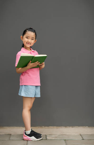Adorable smart primary school student girl smiling looking at camera, sitting on steps at home with a book in hands. Intelligent little girl reading a book. World Book Day concept. Back to school