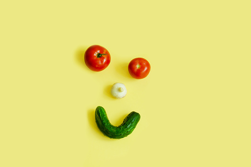 Happy vegetables, vegetable face, tomato eyes, garlic nose, cucumber smile, top view, on a yellow background, copy space. High quality photo
