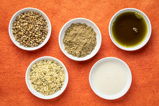 collection of hemp seed products: hearts, protein powder, milk and oil in small white bowls against textured orange paper, superfood concept