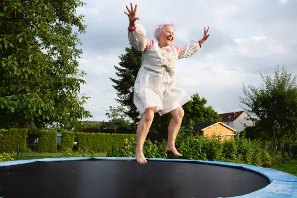 Senior woman with overweight jumping on trampoline Senior woman with dyed pink hair, 67 years old, wearing a white dress with colorful embroidery, jumping on a trampoline in backyard, outdoors at a summer evening, she has overweight and has fun exercising trampoline stock pictures, royalty-free photos & images