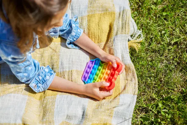 Photo of Girl lies on a blanket on grass outdoors and play pop it, kid hands playing with colorful pop It, fidget toy in the backyard of the house on a sunny summer day, summer time vacation.