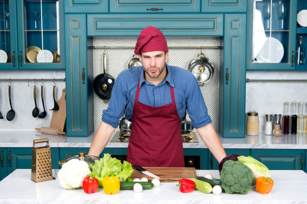 istock Cook stand at kitchen table. Man in chef hat and apron in kitchen. Vegetables and tools ready for cooking dishes. Vegetarian menu and healthy diet. Food preparation and cooking recipes 1329694245