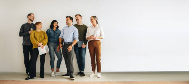 Diverse creative team looking happy Colleagues talking while standing at wall. Happy creative team standing at wall and discussing. colleague stock pictures, royalty-free photos & images