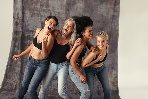 Women of different ages having fun while wearing jeans and bras. Four body positive women smiling cheerfully and celebrating their natural bodies against a studio background.