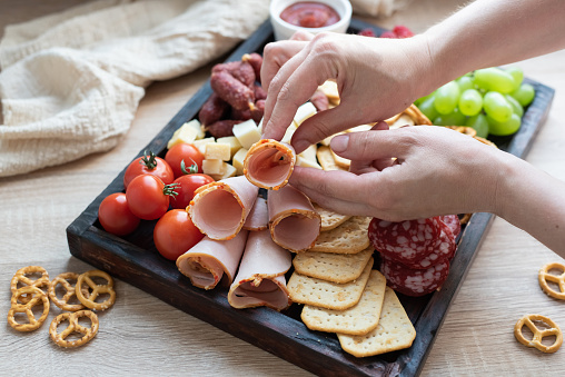 Female hands laying the ham on charcuterie board with sausage, fruit and cheese, party cooking, close-up.