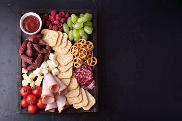 tomates, salchichas, frutas, galletas y queso sobre tabla de charcutería sobre fondo negro, bocadillos de fiesta. - cheese antipasto cracker grape fotografías e imágenes de stock