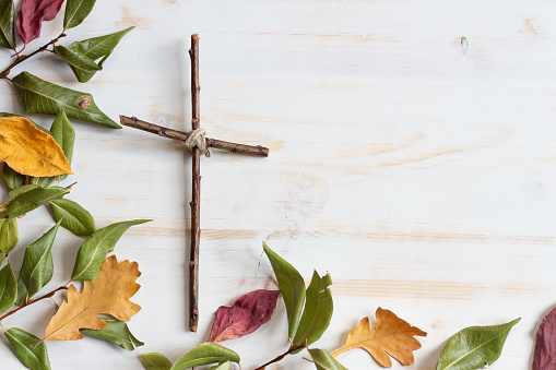 simple wood christian cross with red, yellow and green dried autumn leaves on white wood background with copy space