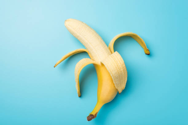 孤立したパステルブルーの背景に真ん中に1つの皮をむいた熟したバナナのトップビュー写真 - 剥いた ストックフォトと画像