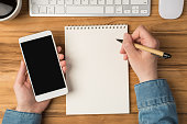First person top view photo of hands holding smartphone and pen over notebook cup of coffee keyboard mouse on isolated wooden desk background with copyspace
