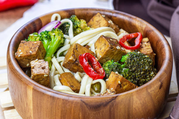 hausgemachter vegetarischer gebratener tofu mit udon-nudeln und gemüse (brokkoli, pfeffer und zwiebeln) in der holzschüssel auf hellem küchenhintergrund - tofu chinese cuisine vegetarian food broccoli stock-fotos und bilder