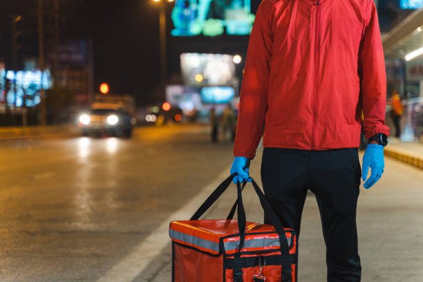 Close-up shot of a food deliveryman in red uniform carrying a food delivery box to deliver for customer for order during COVID-19 outbreak lockdown in the city at night time stock photo