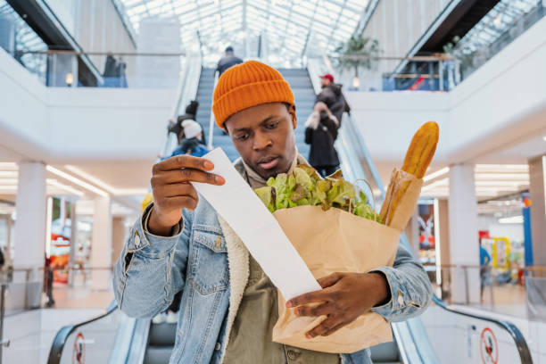 Surprised black man looks at receipt total with food in mall Surprised African-American man in denim jacket looks at receipt total in sales check holding paper bag with products in mall price stock pictures, royalty-free photos & images