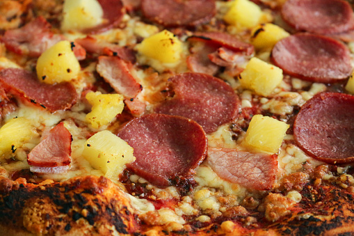 Stock photo showing close-up view of pepperoni pizza topped with a rich tomato sauce, melted grated mozzarella, pineapple pieces, ham and circles of pepperoni sausage.