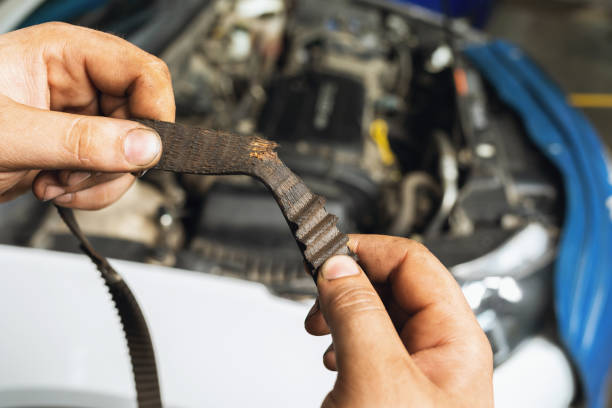 un meccanico automobilistico mostra una cintura di temporizzazione strappata con denti usurati sullo sfondo di un primo piano del cofano dell'auto aperto - deformato foto e immagini stock