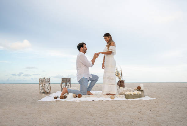 Loving couple getting engaged on a romantic picnic at the beach Loving couple getting engaged on a romantic picnic at the beach and him on his knees while he puts the ring on her finger - relationship concepts fiancé stock pictures, royalty-free photos & images