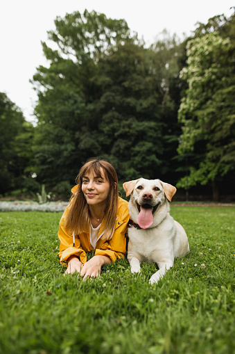 A portrait of a young beautiful woman wearing in yellow rain cover jacket lying and relax with her cute Labrador dog on the grass in public park