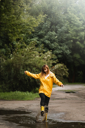 A woman wearing in waterproof yellow jacket and boots standing and jumping on the puddle in the street