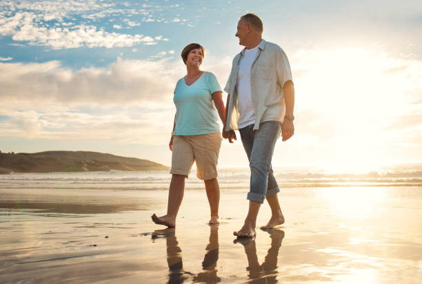 foto de una pareja madura cogiéndose de la mano mientras camina por la playa - pareja madura fotografías e imágenes de stock