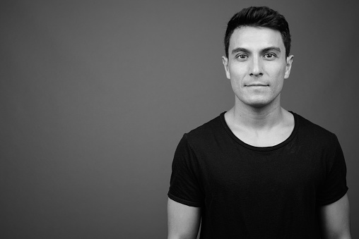 Studio shot of young handsome Hispanic man wearing black shirt against gray background in black and white