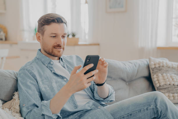 istock Young man sitting on couch and surfing web on his phone 1329727031