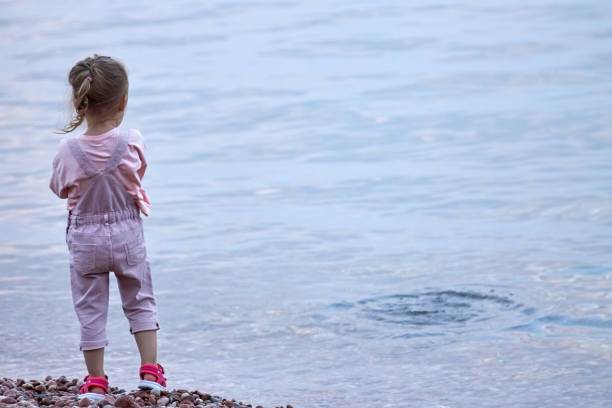 dziewczynka w różowym kombinezonie stoi sama na plaży i patrzy w dal - worry stones zdjęcia i obrazy z banku zdjęć