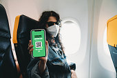 Woman on airplane is wearing a protective face mask and is showing a Covid-19 digital passport to the camera with her smart phone
