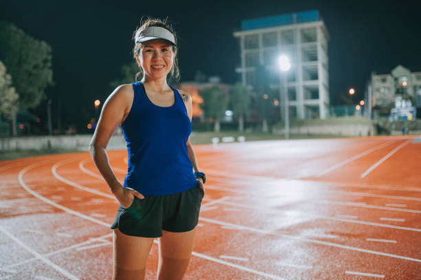 confidence and satisfied female athlete hands in pockets looking at camera standing on all-weather-track and field stadium at night - sports track track and field stadium sport night imagens e fotografias de stock