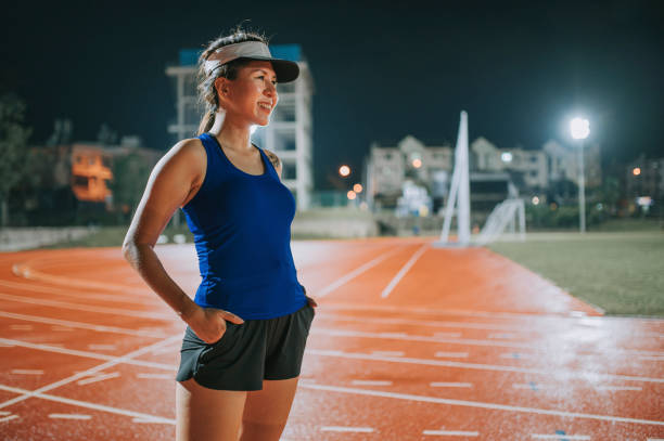 confidence and satisfied female athlete hands in pockets looking at away standing on all-weather-track and field stadium at night - sports track track and field stadium sport night imagens e fotografias de stock