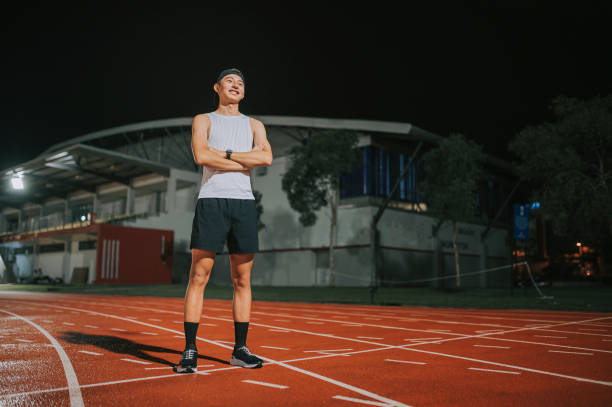 confidence and satisfied male athlete arms crossed looking away standing on all-weather-track and field stadium at night - sports track track and field stadium sport night imagens e fotografias de stock