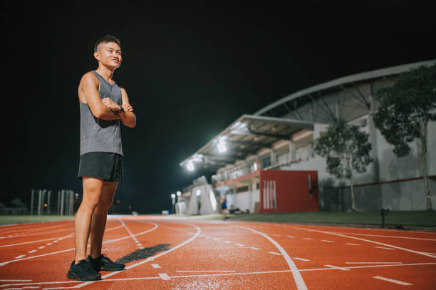 confidence and satisfied male athlete arms crossed looking away standing on all-weather-track and field stadium at night - sports track track and field stadium sport night imagens e fotografias de stock