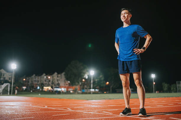 confidence and satisfied male athlete hands on waist arms akimbo looking away standing on all-weather-track and field stadium at night - sports track track and field stadium sport night imagens e fotografias de stock