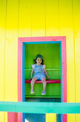 Little girl sitting inside of colorful wooden toy house during summer day