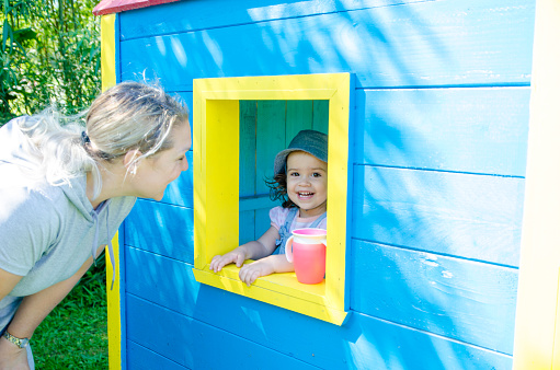 Little girl looking at grandmother out of window from inside of colorful wooden toy house during summer day