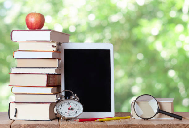istock Stack of books with digital tablet, red apple, silver clock, coloured pencils, magnifying glass in the nature 1329751093