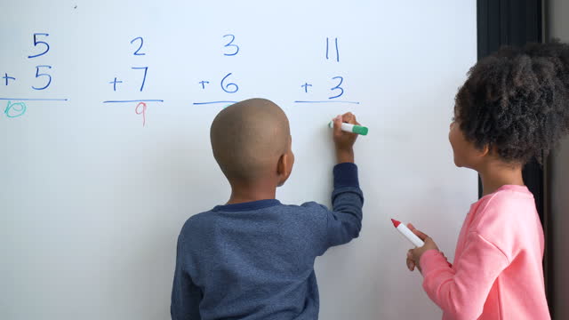 Zwei afroamerikanische Kinder, Geschwister, stehen an einem großen Whiteboard mit Markern, die rechnen. Sie arbeiten zusammen, um Additionsprobleme zu lösen. Der 7-jährige Junge und das 8-jährige Mädchen zählen an ihren Fingern. Sie reden darüber, und dann schreibt der Junge die Antwort. Sie geben sich gegenseitig High Fives.