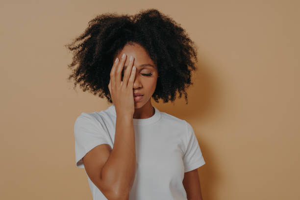 mujer de piel oscura cubriendo la cara con la mano y sintiendo ansiedad o depresión contra la pared beige - estrés fotografías e imágenes de stock