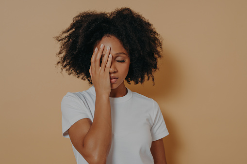 Studio shot of young upset mixed race woman being frustrated by problems in life or relationships, covering face with hand and feeling anxiety or depression while posing against beige wall in studio
