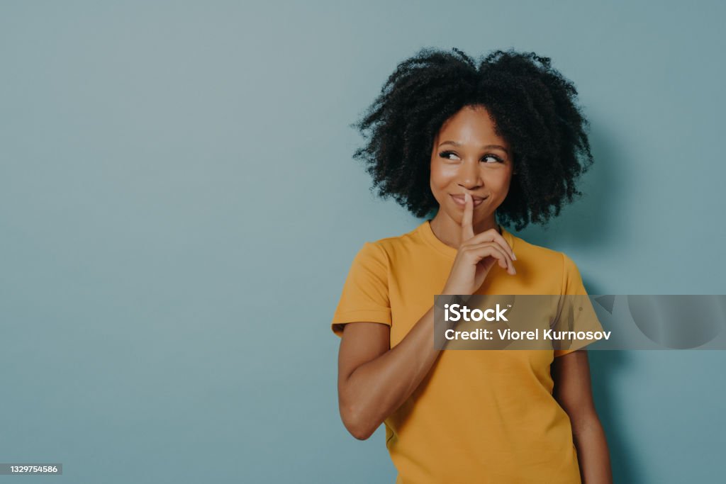 Cheerful afro girl showing shhh sign with finger near lips, standing over pastel blue background Keep silence gesture. Cheerful afro girl showing shhh sign with finger near lips, standing over pastel blue background with copy space and mysteriously looking away. Positive women emotions concept Privacy Stock Photo