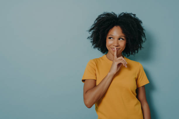 Cheerful afro girl showing shhh sign with finger near lips, standing over pastel blue background Keep silence gesture. Cheerful afro girl showing shhh sign with finger near lips, standing over pastel blue background with copy space and mysteriously looking away. Positive women emotions concept mystery stock pictures, royalty-free photos & images