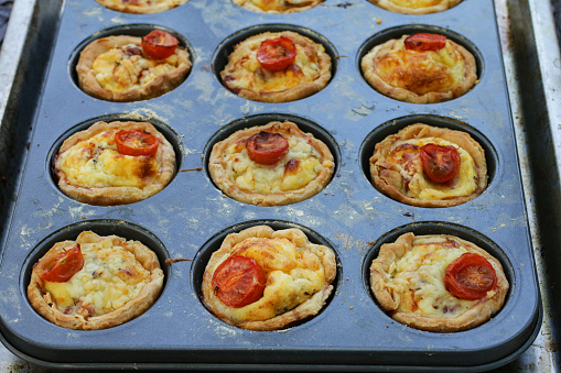 Stock photo showing a close-up view of baking tray full of individual, mini, golden, bacon quiches pies left to cool in baking tin, on a kitchen worktop, after being removed from the oven.