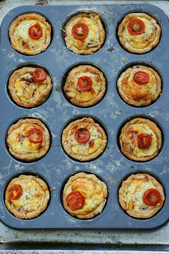 Stock photo showing an elevated view of baking tray full of individual, mini, golden, bacon quiches pies left to cool in baking tin, on a kitchen worktop, after being removed from the oven.