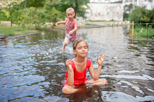 Cute girl sitting in lotus pose in puddle after warm summer rain and her brother jumping and swimming around her, outdoor walking in any weather, happy childhood and summertime
