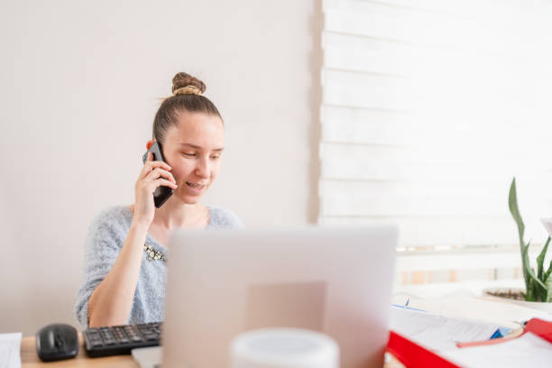 A businesswoman is working at home and talking on a smart phone while using a laptop.